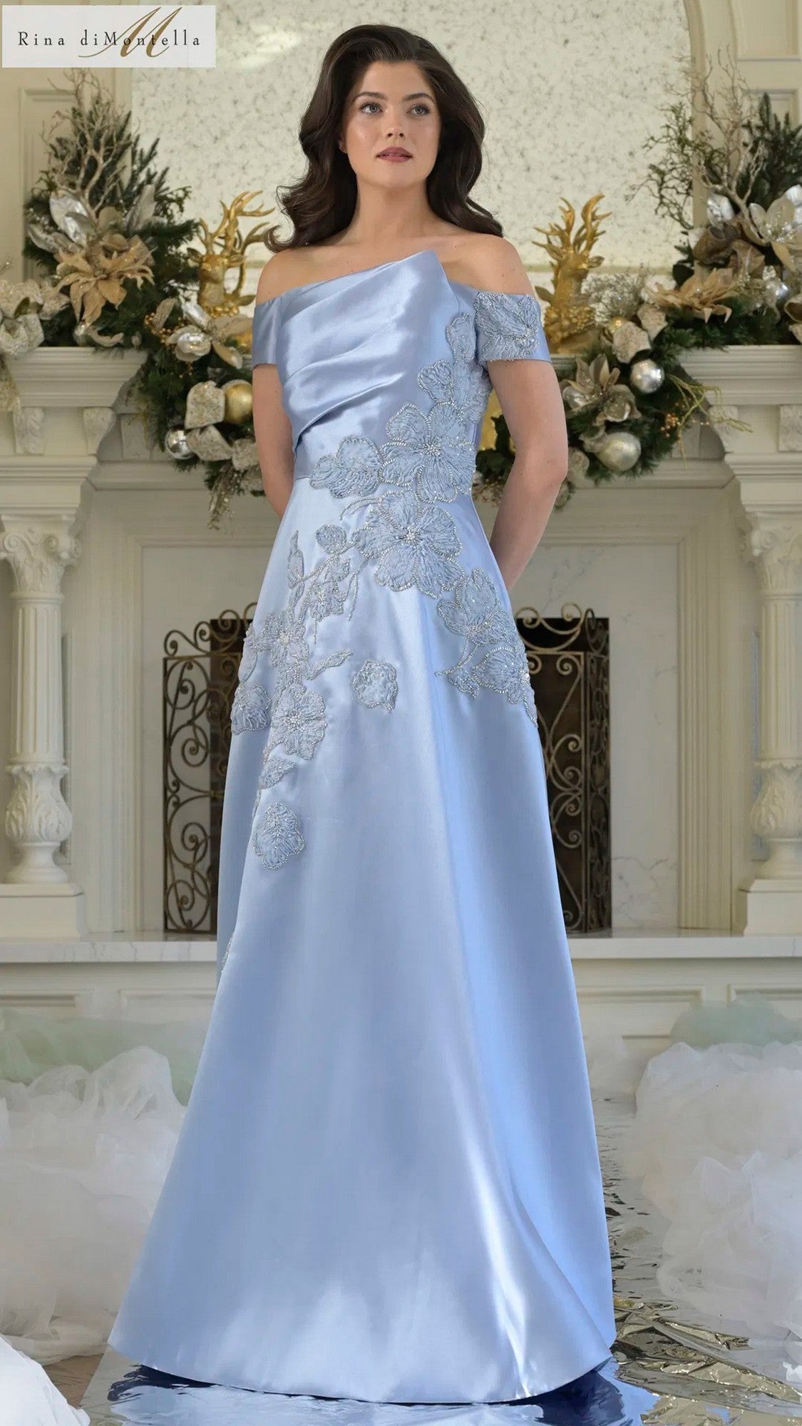 A woman stands in a sky blue Rina Di Montella RD3344 long formal dress with silver floral embroidery, posed before an ornate white mantel decorated with gold and white floral arrangements.